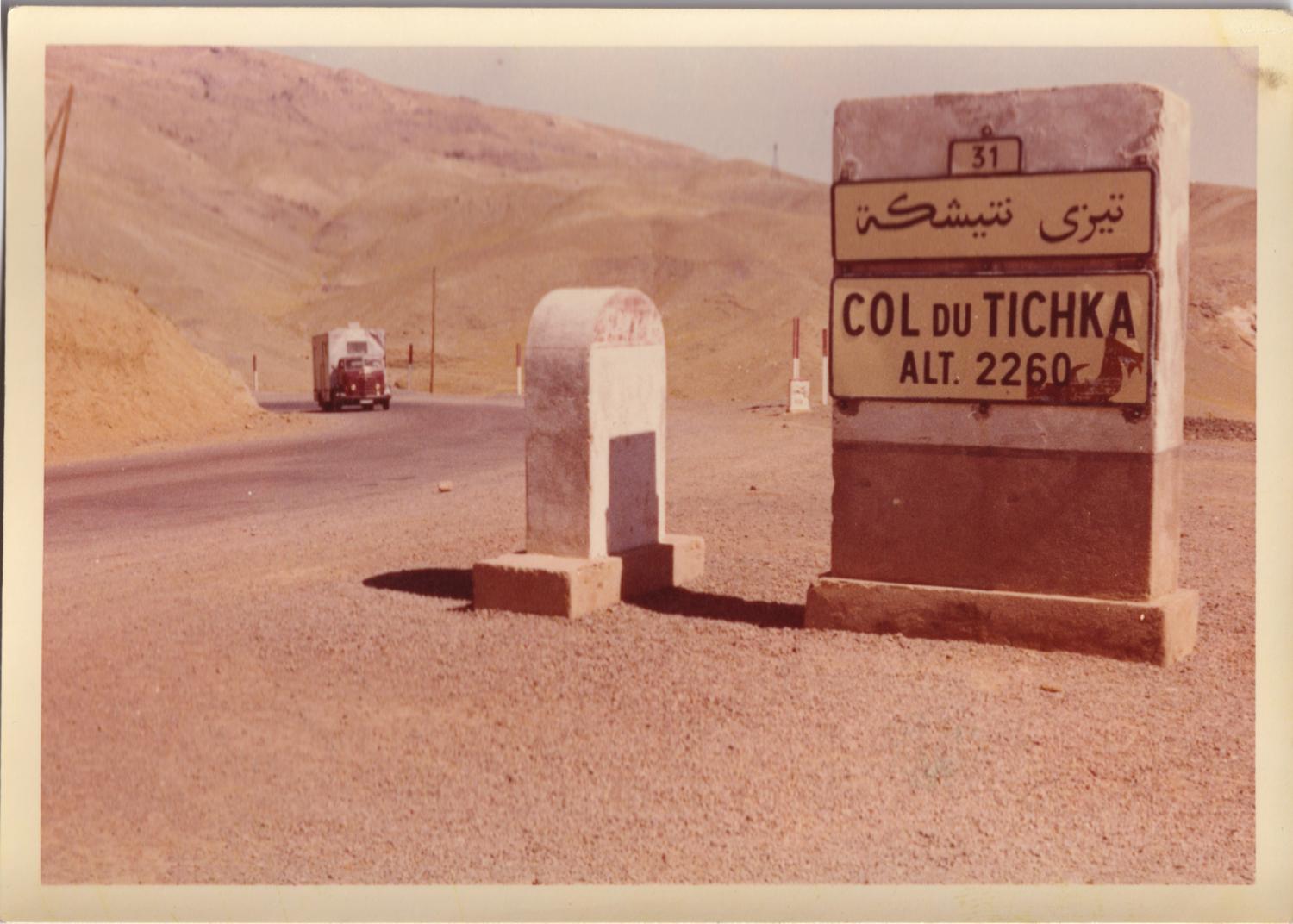 A roadside marker in the High Atlas reading Col du Tichka, altitude 2260, in French and Arabic — with a catering van coming up the pass behind it