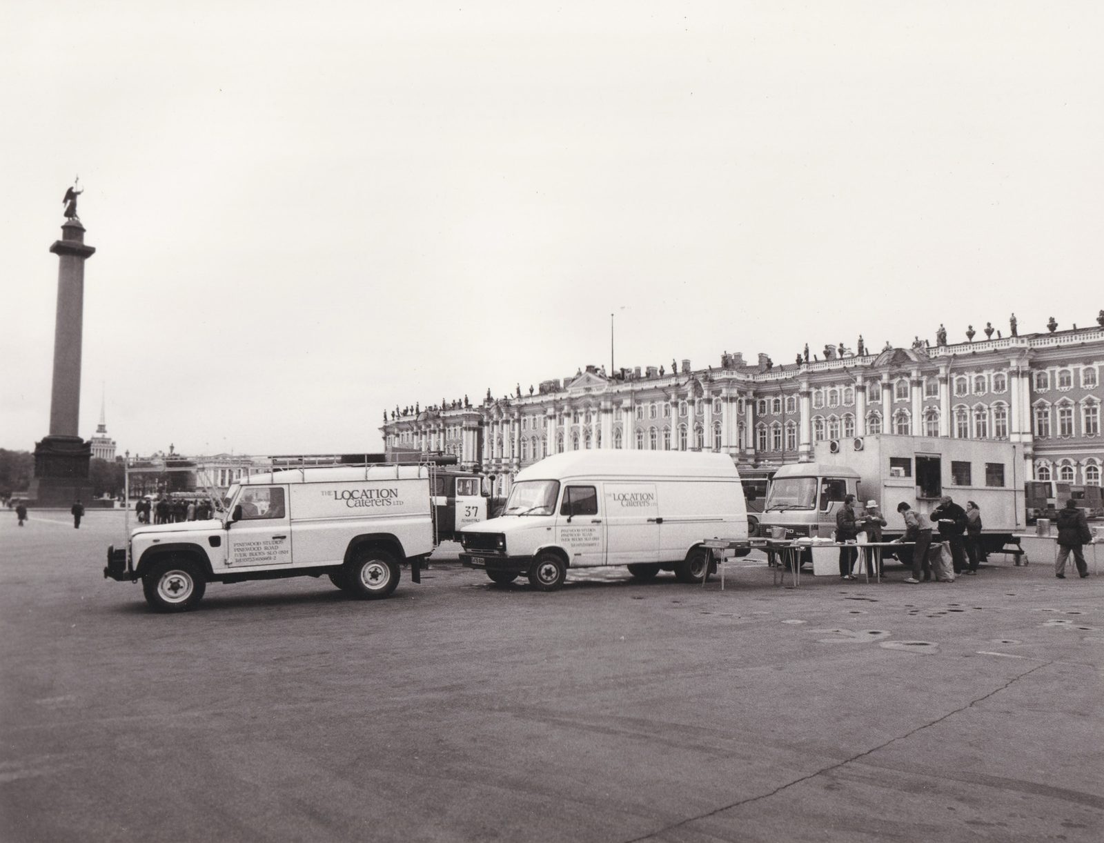 Two vans lettered The Location Caterers Ltd, Pinewood Studios, parked on the cobbles of Palace Square in Leningrad with the Winter Palace behind them and the Alexander Column to the left. Beside a catering truck at the right, a film unit is being served lunch at trestle tables.